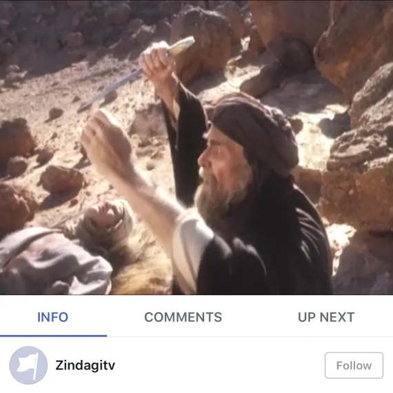 Man in traditional clothing kneeling beside another man lying on rocky ground, raising hand in distress.