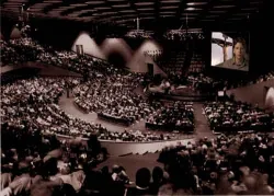 Large auditorium filled with people, speaker shown on a screen above the stage.