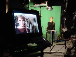 Woman sitting in front of a green screen with her image on a monitor nearby.